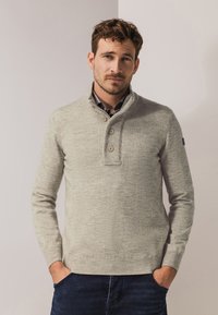 Hellegrauer Pullover mit hohem Kragen, einer drei-Knopf-Leiste und gerippten Bündchen sowie Saum. Über einem karierten Hemd getragen.