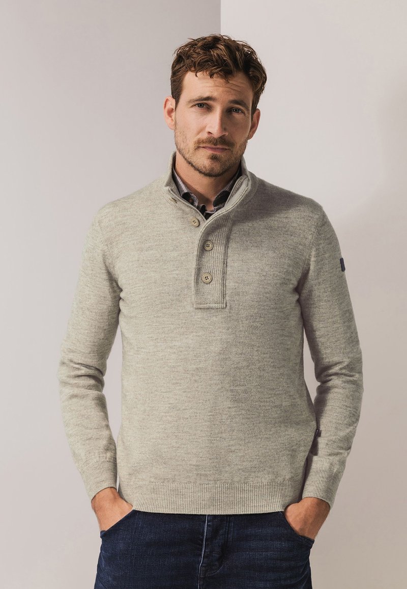 Hellegrauer Pullover mit hohem Kragen, einer drei-Knopf-Leiste und gerippten Bündchen sowie Saum. Über einem karierten Hemd getragen.