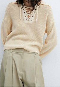Beige knitted sweater with a lace-up neckline, featuring eyelets and a relaxed fit. Paired with light beige high-waisted trousers.