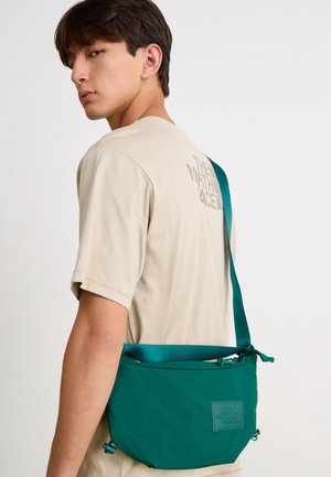 Ung mand iført en beige The North Face T-shirt og en grøn The North Face crossbody-taske på ryggen.