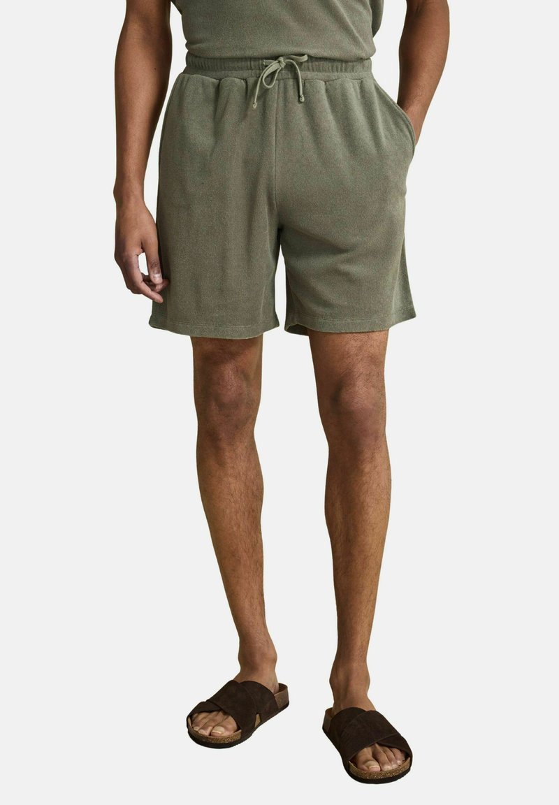 Homme portant un short à cordon de serrage vert olive avec poches latérales et des sandales marron glissantes, debout devant un fond uni.