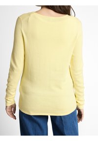Femme portant un pull uni jaune pâle à manches longues et un jean bleu, vue de dos sur fond blanc.