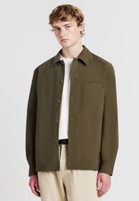 Camicia verde oliva con bottoni, caratterizzata da una tasca sul petto, realizzata in tessuto morbido e con colletto classico e maniche lunghe.