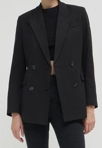 Blazer noir à double boutonnage en tissu lisse, avec des revers en pointe, deux poches avant et quatre boutons noirs sur le devant.