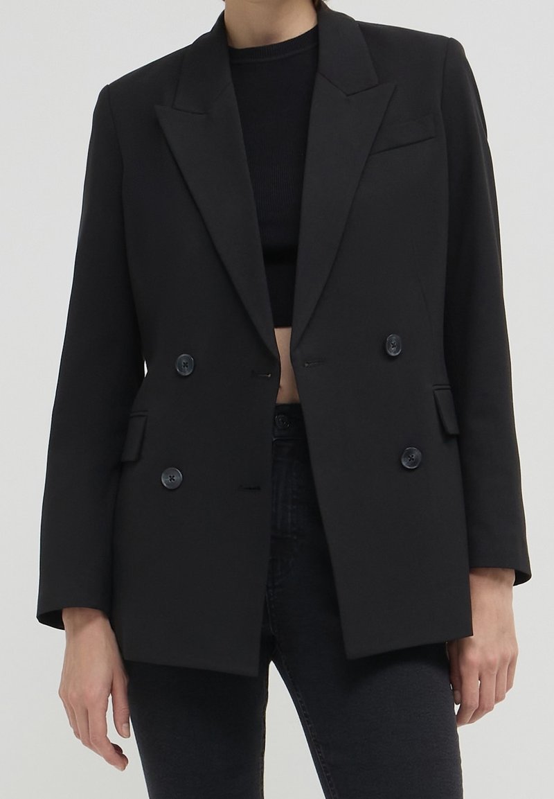 Blazer noir à double boutonnage en tissu lisse, avec des revers en pointe, deux poches avant et quatre boutons noirs sur le devant.