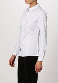 Persona che indossa una camicia bianca a maniche lunghe aderente con bottoni e pantaloni neri, in piedi davanti a uno sfondo semplice.