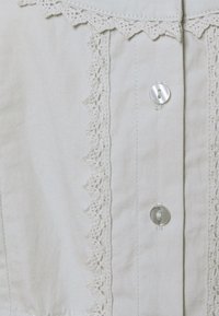 Chemise en tissu gris clair avec un galon en dentelle détaillé le long de l'encolure et une rangée de boutons transparents sur le devant. Texture douce.