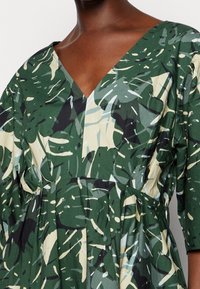 Femme portant une robe verte avec un imprimé feuillage abstrait, un décolleté en V et des manches 3/4 légèrement bouffantes.