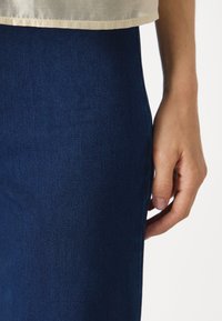 Dark blue denim skirt with a smooth texture and tailored fit, featuring a side seam and a hand resting on the fabric.