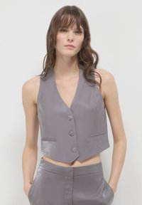 Gilet gris sans manches avec un col en V, texture brillante, trois boutons à l'avant et deux poches latérales, associé à un pantalon assorti.