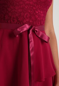 Robe bordeaux avec un corsage en dentelle, une ceinture en ruban de satin et une jupe en mousseline à couches avec un ourlet asymétrique. Texture douce dans l'ensemble.