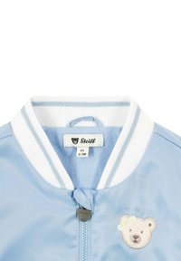 Hellblaues Bomberjacke mit weißem gestreiftem Kragen, versehen mit einer Bärenstickerei auf der Brust und einem Reißverschluss mit einem bärenförmigen Zipper. Größenetikett sichtbar.