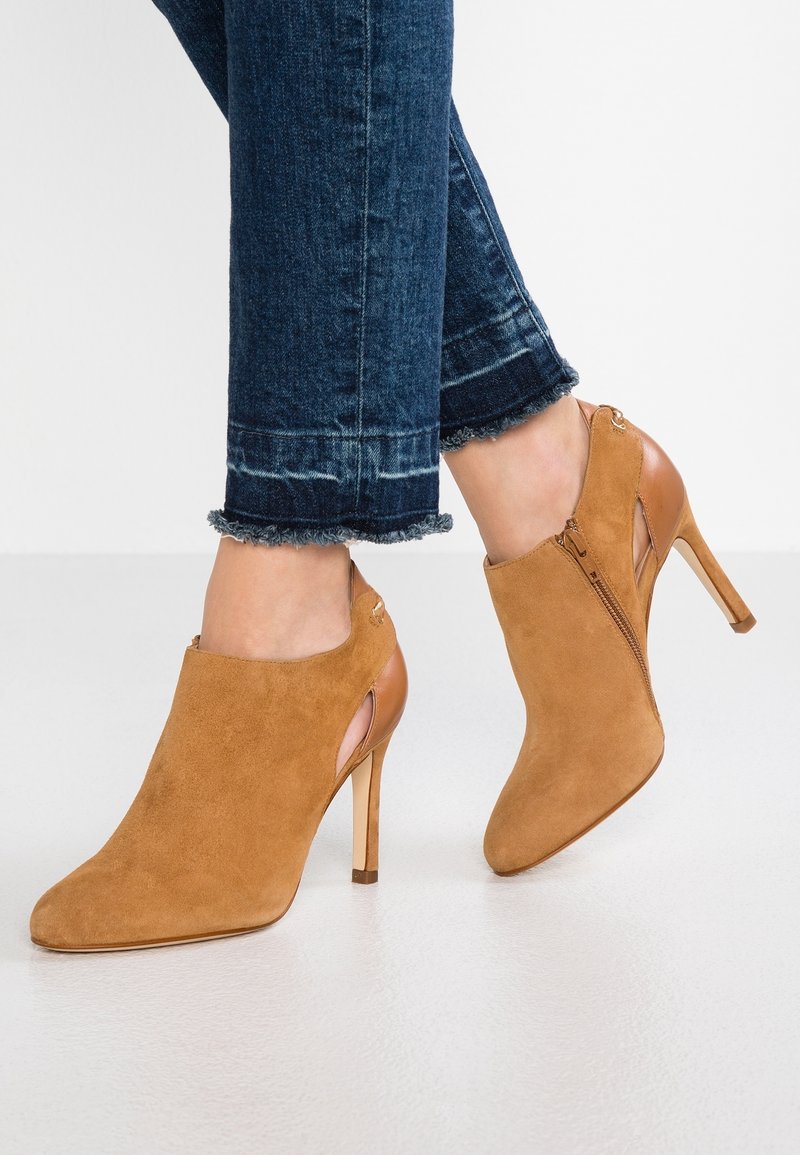 Pieds portant des bottines à talons hauts en daim beige avec des découpes latérales et des fermetures éclair, associées à un jean bleu foncé à ourlets effilochés.