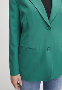 Blazer verde entallado con dos botones, solapas notch, bolsillos laterales y tela suave. Combinado con jeans de mezclilla azul.