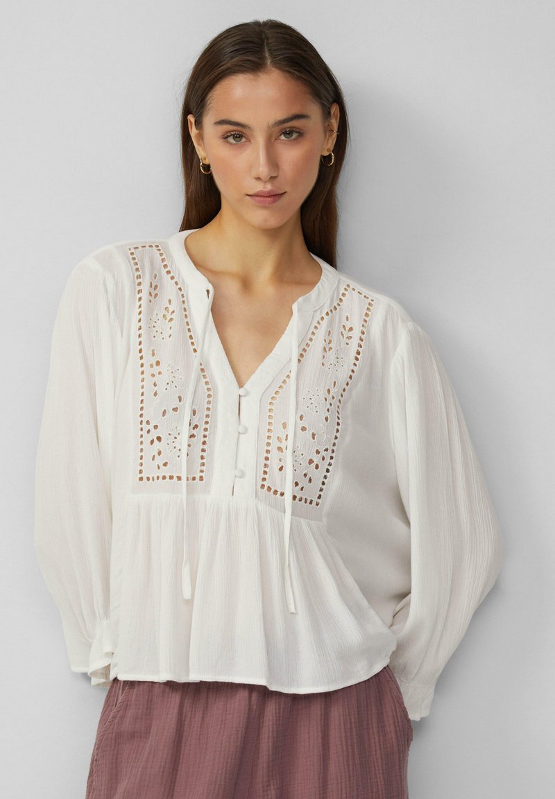 Weiße Bluse mit V-Ausschnitt, filigraner Stickerei, Knöpfen vorne und fließenden Ärmeln; hat einen gerafften Saum.