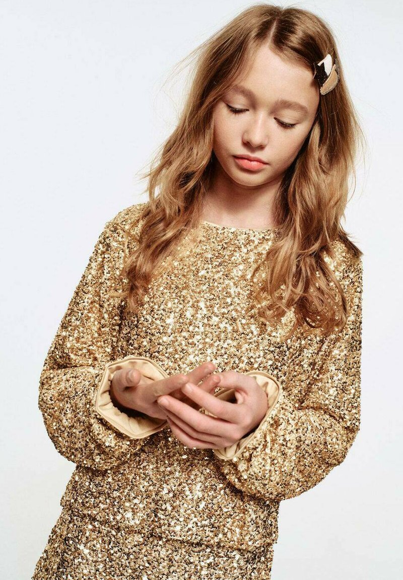 Maglione oro con paillettes, maniche lunghe e polsini crema arricciati; fermacapelli color marrone chiaro; modello con le mani giunte davanti.