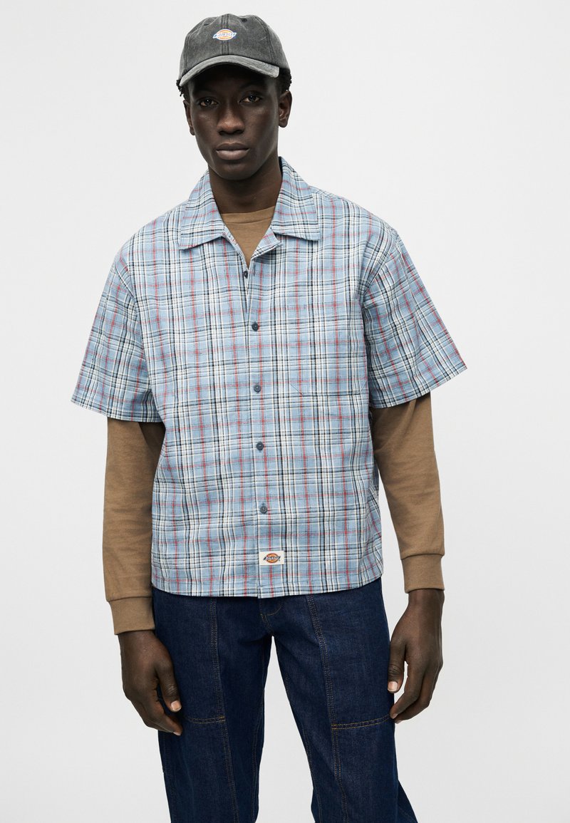 Junger Mann mit grauer Mütze, blau-rotem kariertem Kurzarmhemd über einem braunen Langarmshirt und dunkelblauen Jeans.