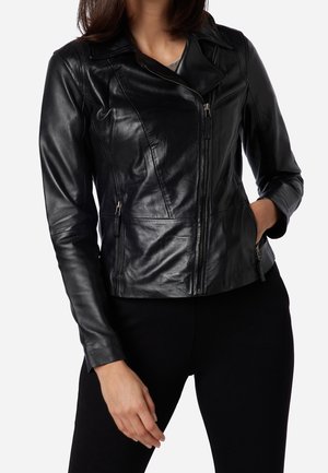 Kvinde iført tætsiddende sort læderjakke med lynlås foran, lynlåse i sidelommer og asymmetrisk krave, kombineret med sorte bukser.