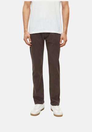 Mann trägt dunkelbraune gerade Hose, weiße Sneaker und ein weißes T-Shirt und steht vor einem schlichten Hintergrund.