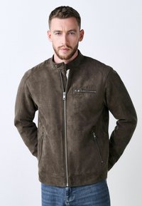 Chaqueta de ante marrón con cuello alto, cierre frontal con cremallera y dos bolsillos laterales. Cuenta con un bolsillo en el pecho con cremallera y una textura suave.