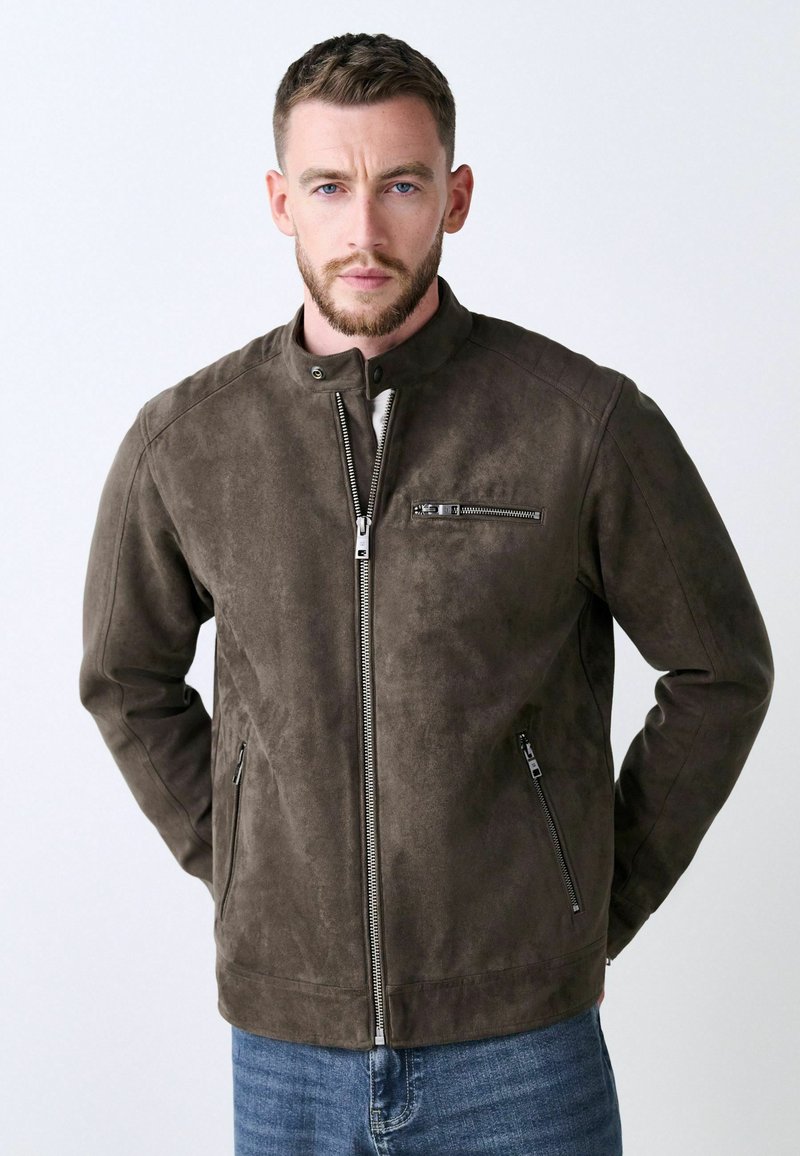 Chaqueta de ante marrón con cuello alto, cierre frontal con cremallera y dos bolsillos laterales. Cuenta con un bolsillo en el pecho con cremallera y una textura suave.