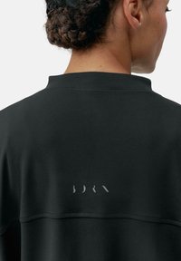 Must mustasvärk, millel on kõrge kaelus ja sile tekstuur, ning mille taga kaeluses on peen hall logo.