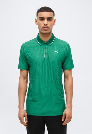 Mann trägt ein grünes Puma-Poloshirt mit dezenten Mustern und einem weißen Puma-Logo auf der Brust, steht vor einem schlichten Hintergrund.