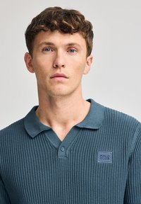 Camisa polo de canalé en verde oscuro con una tapeta de dos botones, cuello corto y un parche con un logo cuadrado en el lado izquierdo del pecho.
