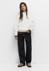 Hvid hættejakke med pink grafik, lagdelt over en brun skjorte. Sorte vidde jeans og beige fodtøj med pink snørrebånd. Afslappet outfit.