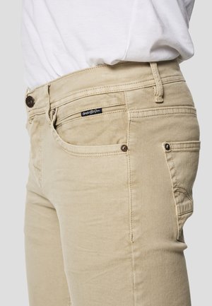 Smėlio spalvos medvilninės kelnės su prigludusiu siluetu, priekyje kišenės, užtrauktuko užsegimas ir mažas prekės ženklo lipdukas ant liemens.