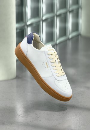 Weißer Hugo-Sneaker mit beigen Schnürsenkeln, brauner Sohle und marineblauem Fersendetail, schwebt über poliertem Betontboden in der Nähe von Glasbausteinen.