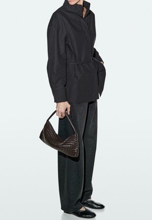 Žena v černém kabátu s páskem, širokých černých kalhotách, černých balerínách a držící po straně tkanou tmavě hnědou kabelku.