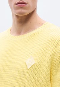 Κίτρινο πουλόβερ  πλεκτό με λευκό μπαλώμα σε σχήμα διαμαντιού. Διαθέτει ριμπ κορυφή και υφασμάτινη υφή. Περιλαμβάνεται λεπτομέρεια στον ώμο.