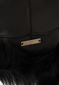 Acercamiento a un sombrero de cuero negro y pelo con costuras visibles y una pequeña etiqueta metálica rectangular unida con hilo negro.