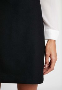Gonna nera in tessuto morbido. La mano di una modella poggia sul bordo, mettendo in mostra il design sartoriale del capo e le sue linee pulite.