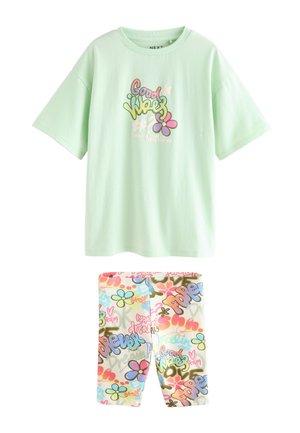 Blondes Mädchen mit offenem Haar trägt ein mintgrünes oversized "Good Vibes" T-Shirt und bunte gemusterte Shorts, das vor einem beigen Hintergrund steht.