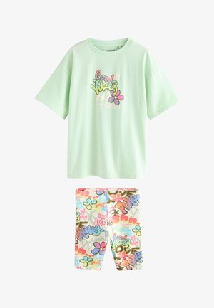 Blondes Mädchen mit offenem Haar trägt ein mintgrünes oversized "Good Vibes" T-Shirt und bunte gemusterte Shorts, das vor einem beigen Hintergrund steht.