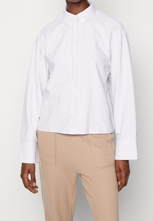 Henkilö, joka käyttää valkoista ja vaaleanharmaata pystysuoraa raidallista pitkähihnaista paitaa, jossa on ylisuuret kalvosimet, ja beigejä housuja, joissa on etutaskut.