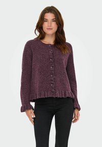 Cardigan violet, texturé, avec un col rond, fermeture à boutons et ourlet froncé. Manches longues avec des poignets froncés. Porté sur un pantalon noir.