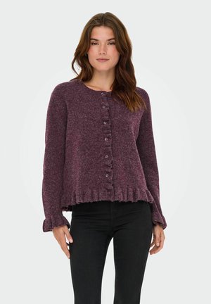 Cardigan violet, texturé, avec un col rond, fermeture à boutons et ourlet froncé. Manches longues avec des poignets froncés. Porté sur un pantalon noir.