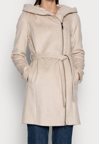 Abrigo beige con cinturón en un material suave y texturizado, con una gran capucha, cremallera frontal completa y bolsillos laterales. Largo hasta mitad del muslo, con un corte entallado.