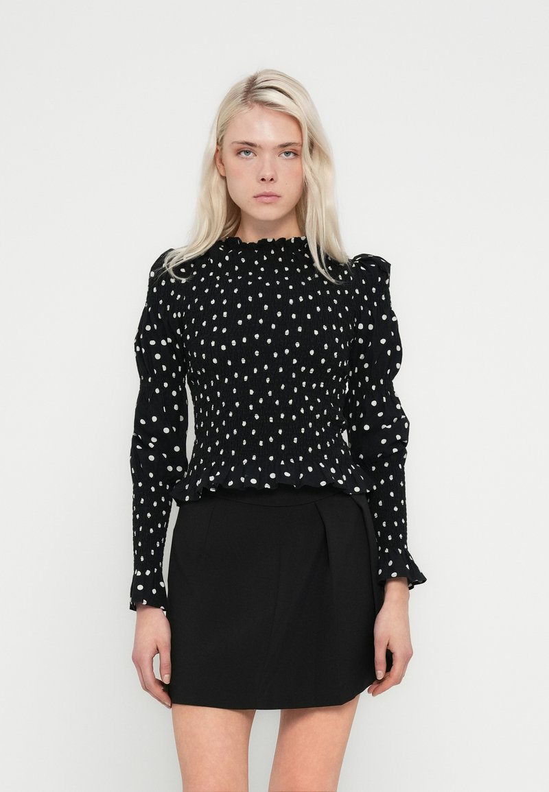 Bluza neagră cu mâneci lungi, cu buline albe, textură cu șnururi elastice, cu guler și manșete volanate. Asortată cu o fustă neagră simplă.