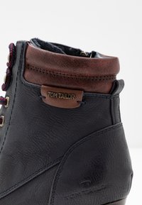 Botín negro de piel con un acento marrón, diseño con cordones, acabado texturizado y una etiqueta de logotipo metálico. Presenta costuras y ojales duraderos.