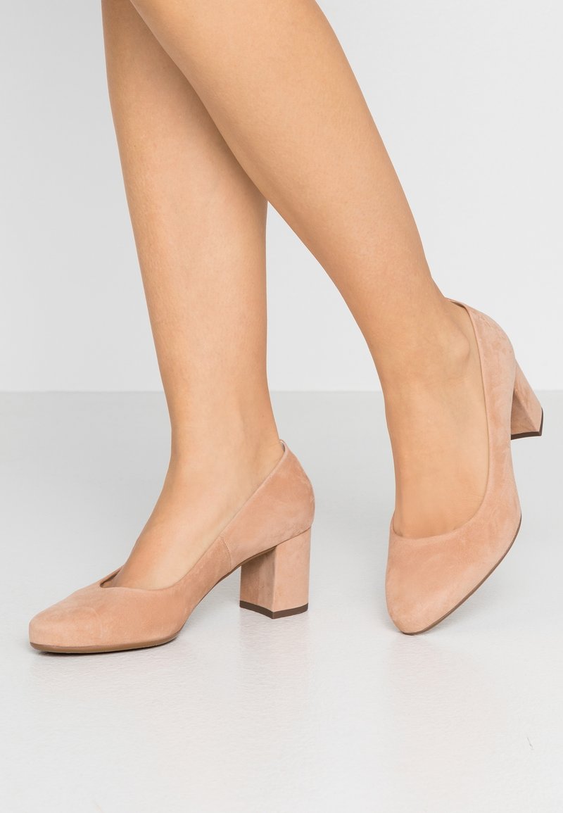 Jambes portant des escarpins à talons carrés en daim couleur nude sur un sol lisse gris clair, sur un fond uni clair.