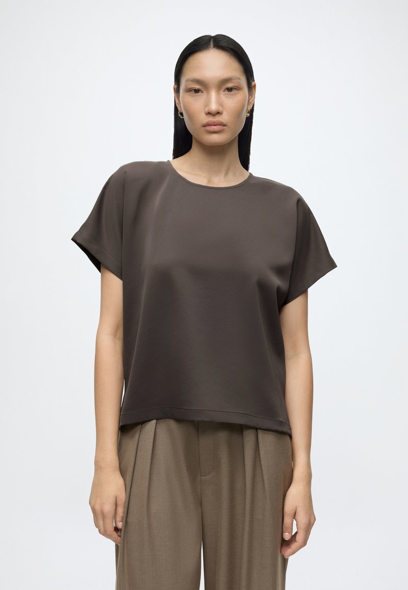 Femme aux cheveux noirs et raides portant un haut ample à manches courtes marron foncé et un pantalon plissé marron clair, debout devant un fond uni.