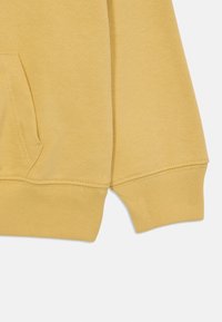 Manica di felpa gialla in cotone con polsino a coste e dettaglio della tasca laterale. Tessuto liscio con cuciture sottili attorno all'orlo.