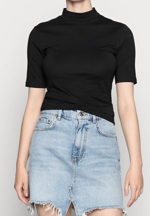 Person iført en tætsiddende sort top med korte ærmer, der er stoppet ned i et lyseblåt højtaljet denimskørt med en frontslids og frynset kant.