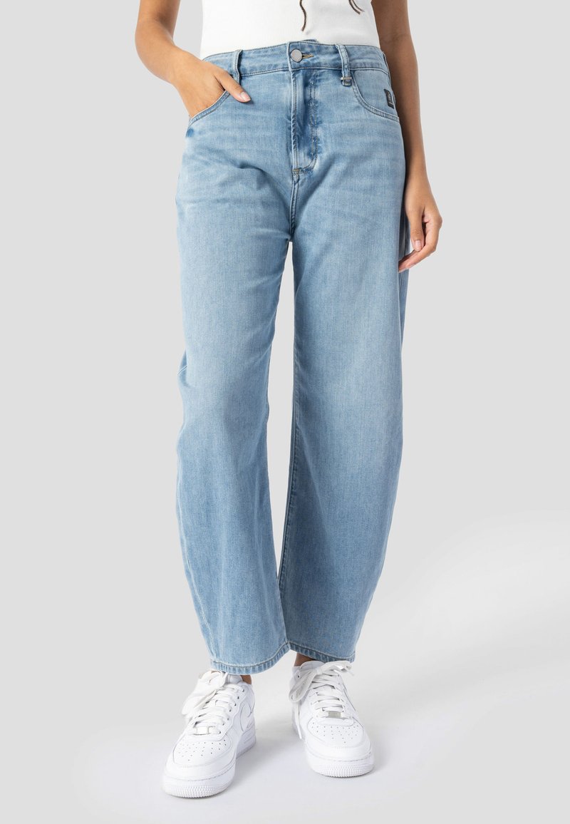 Woman wearing light blue straight-leg jeans and white sneakers with hands in pockets, standing against a plain background.