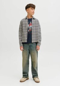 Un garçon se tenant debout, portant des chaussures beiges, un jean large délavé, un t-shirt graphique bleu marine et une chemise à carreaux ouverte, regardant vers la gauche.