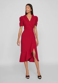 Piros, térdig érő ruha pöttyös ujjakkal, mély V-nyakkal és átlapolt stílussal, fodros szegéllyel és oldalsó sliccsel. Fekete magassarkúval párosítva.